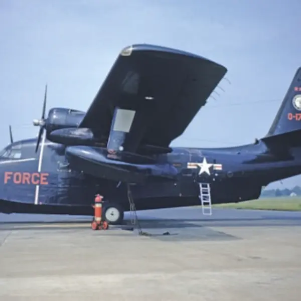 A dark-colored military aircraft marked with 'U.S. Air Force' in bold letters on its fuselage, stationed on an airfield. An aerial ladder is propped against the side of the aircraft near the wing. A red and white fire hydrant can be seen in front of the aircraft's nose. The background shows a grassy landscape under a clear blue sky.